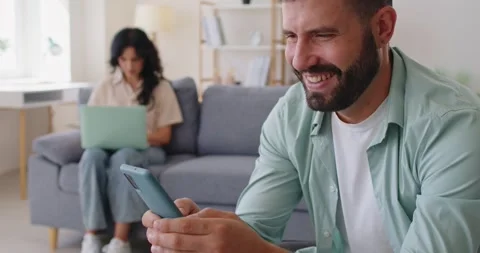 Smiling man using smartphone, partner working behind at home, jealousy in couple Stock Footage 320322155