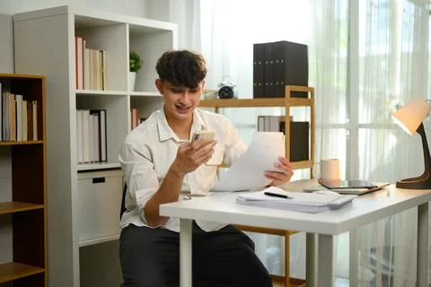 Smiling man using smartphone while reviewing documents in a home office Foto stock
