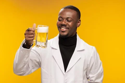 Smiling man in a white lab coat holding a mug of beer against a warm yellow Stock Photos