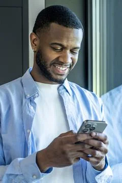 Smiling man by a window in a light blue shirt reads his smartphone, feeling.. Stock Photos