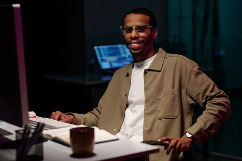 Smiling Man Working at Computer in Dark Office Setting Foto stock