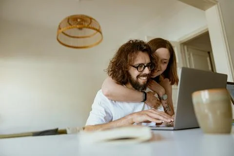 Smiling man working on laptop while wife is hugging him at home. Distance work Stock Photos