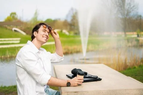 Smiling man working remotely on a laptop in a sunlit park by a fountain. Co.. Stock Photos