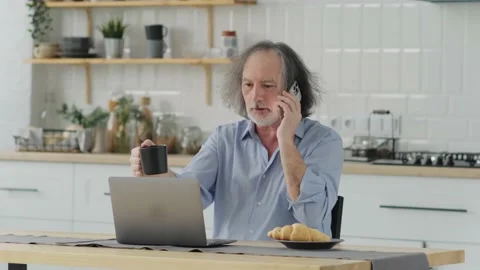 Smiling mature man sit at table at home talk on cellphone work on computer Stock Footage 237628256