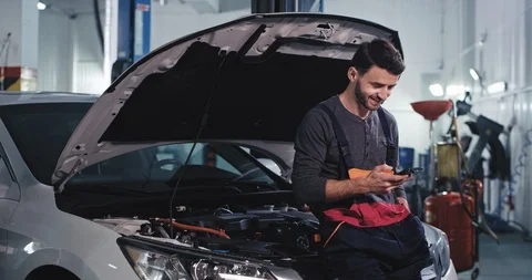 Smiling mechanic guy in a uniform take a look on his phone while fixing the car Stock Footage 120007043