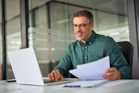 Smiling middle aged executive holding documents using laptop in office at desk. Stock-Fotos