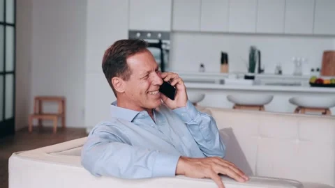 Smiling middle-aged man 60s in blue shirt talking by the phone at home. Sitting Stock Footage 231703595
