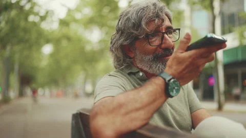 Smiling middle-aged man with gray hair and beard sits on bench and uses mobile Stock Footage 227030186