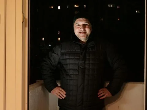 Smiling middle aged man standing on a balcony at night with hands on hips, lo Stock Photos