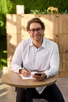 Smiling middle aged man in white shirt sits at round table writing on paper Stock-Fotos