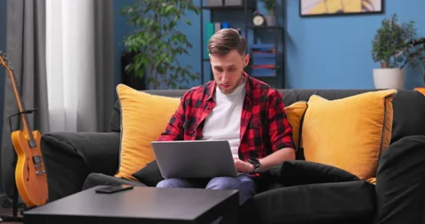 Smiling millennial boy sit relax on couch using modern laptop browsing unlimited Stock Footage 154043436
