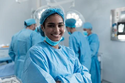 Smiling mixed race female surgeon with face mask and protective clothing in Stock Photos