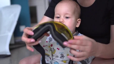 Smiling mother puts on ski goggles on baby with surprised face and eyes wide Видео 80286463