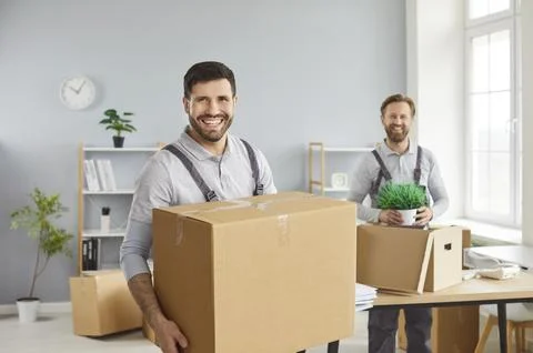 Smiling movers handling cardboard boxes while relocating items in a sunny office Stock Photos