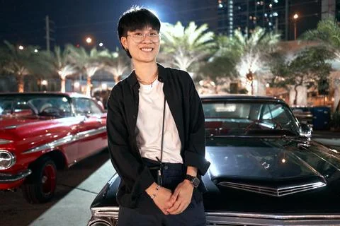 Smiling non-binary person leaning on a luxury car the lot of a night fair Stock Photos