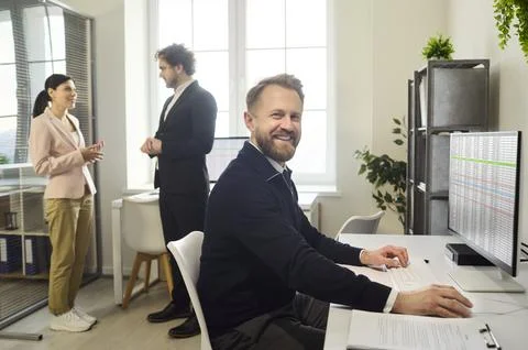 Smiling office employee at computer, colleagues in background, confident Stock Photos