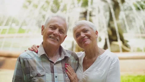 Smiling old couple looking at camera while standing next to a fountain in a park Stock Footage 198716895