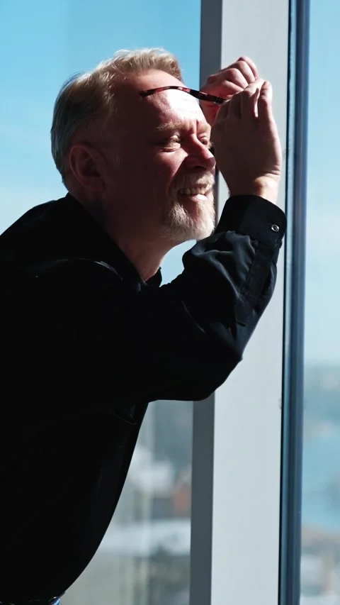 Smiling old man leaning on the window takes off his glasses. Side view of a man Stock Footage 339504621