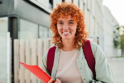 Smiling redhead student looking at the camera while holding a red folder and Stock Photos