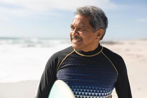 Smiling retired biracial senior man looking away while standing with surfboard Stock Photos