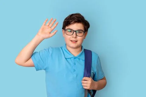 A smiling schoolboy in glasses, with a backpack, waves his hand affably, looks Stock Photos
