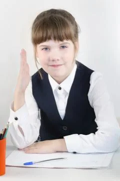Smiling schoolgirl in uniform sit at table with exercise book and raises her  Stock Photos
