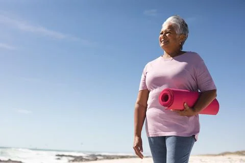 Smiling senior biracial woman holding pink yoga mat while looking away at beach Stock Photos