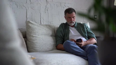 Smiling senior grey-haired man sitting on couch and using smartphone at home Stock Footage 158836759