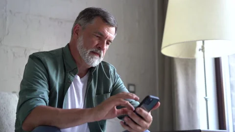 Smiling senior grey-haired man sitting on couch and using smartphone at home Stock Footage 158837161