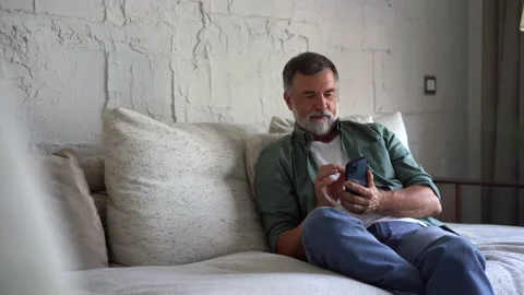 Smiling senior grey-haired man sitting on couch and using smartphone at home Stock Footage 158837177