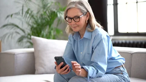 Smiling senior lady using smartphone texting message sit on couch at home Stock Footage 182294071
