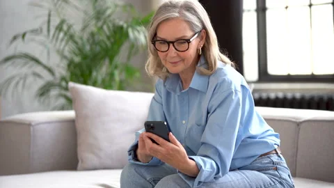 Smiling senior lady using smartphone texting message sit on couch at home Stock Footage 182297645