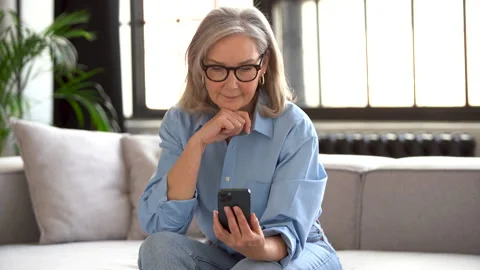 Smiling senior lady using smartphone texting message sit on couch at home Stock Footage 182300927