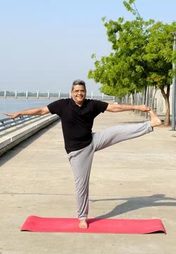 Smiling senior man performing standing leg stretch yoga pose outdoors on a mat Stock Photos