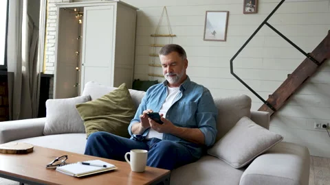 Smiling senior man sitting on couch and using mobile phone at home, chatting Stock Footage 222111617
