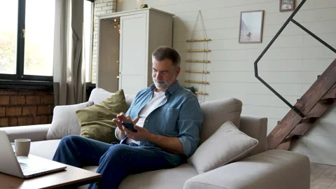 Smiling senior man sitting on couch and using mobile phone at home, chatting Stock Footage 222111798