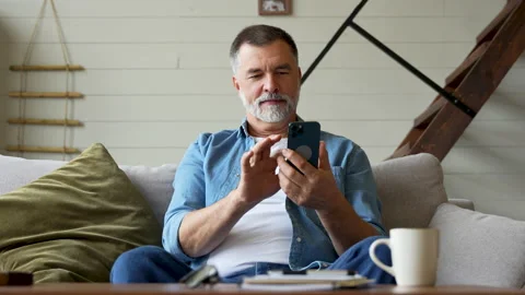 Smiling senior man sitting on couch and using mobile phone at home, chatting Stock Footage 222113814