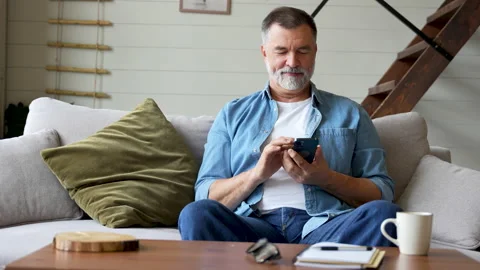Smiling senior man sitting on couch and using mobile phone at home, chatting Stock Footage 222114009