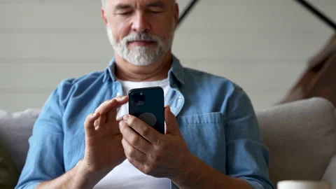 Smiling senior man sitting on couch and using mobile phone at home, chatting Stock Footage 222115926
