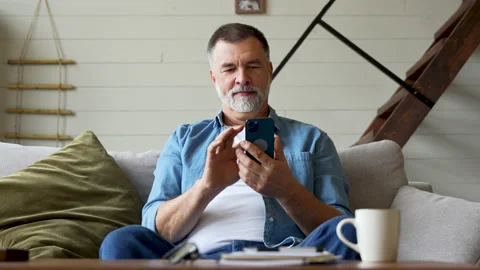 Smiling senior man sitting on couch and using mobile phone at home, chatting Stock Footage 222116438