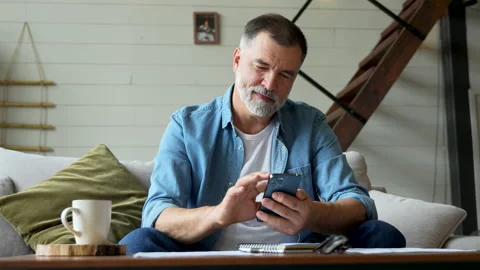 Smiling senior man sitting on couch and using mobile phone at home, chatting Stock Footage 222118162