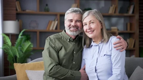 Smiling senior married couple embracing while sitting on a sofa and looking  Stock Footage 339775992