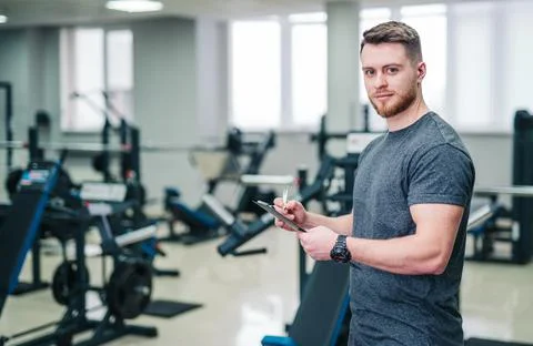 Smiling sportsman standing in light gym with sport equipment and writing. Stock Photos