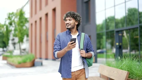 Smiling student with a backpack uses a mobile phone while walking in the campus  Stock Footage 317711057