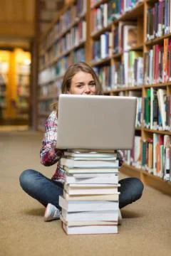 Smiling student sitting on library floor using laptop on pile of books 스톡 사진