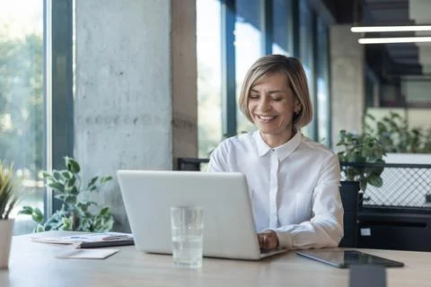 Smiling stylish mature middle aged woman sits at desk with laptop, sitting .. Stock Photos