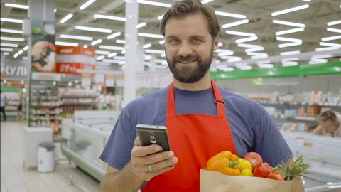Smiling supermarket employee using mobile phone and holding shopping bag in Stock Footage 92237597