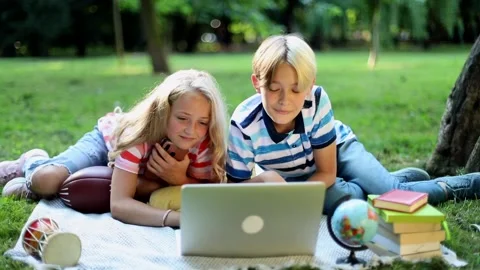 Smiling talking kids using laptop resting in sunny green park outside. Learning  스톡 동영상 137371176