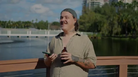 Smiling tattooed man standing on a lakeside boardwalk, Vidéo 323733010