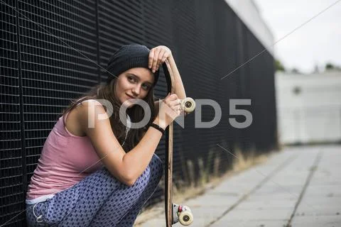 Smiling teenage girl with skateboard crouching in front of a black facade Stock Photos
